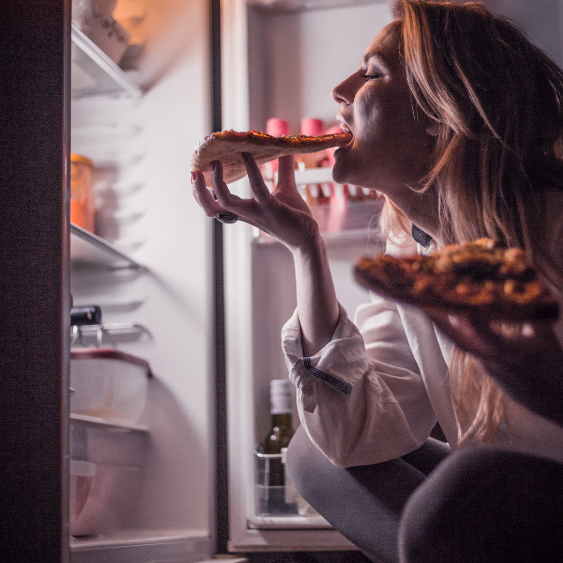 A woman sitting on the floor in front of her refrigerator at night eating pizza.